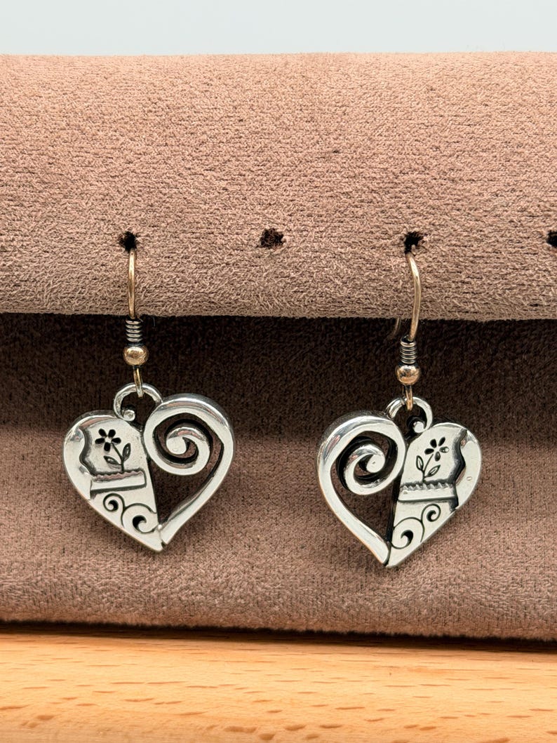May include: A pair of silver-coloured heart-shaped earrings. Each earring has a floral design on one side and a swirling design on the other. The earrings hang from a gold-coloured hook.