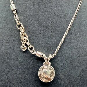 May include: A silver-toned necklace featuring a twisted chain and a round pendant with a woven texture and a central heart motif. The necklace includes an adjustable link section with a small heart-shaped charm bearing the letter B.