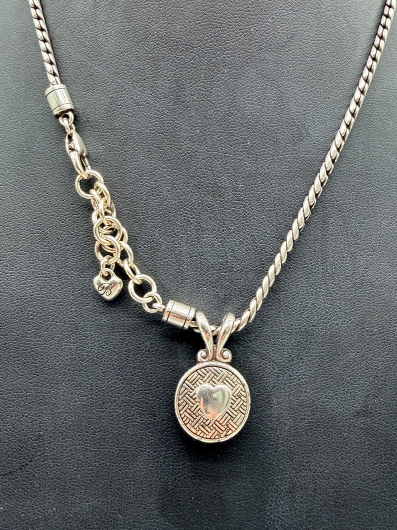 May include: A silver-toned necklace featuring a twisted chain and a round pendant with a woven texture and a central heart motif. The necklace includes an adjustable link section with a small heart-shaped charm bearing the letter B.