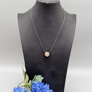 Puede incluir: Un collar plateado con un colgante redondo. El colgante tiene un borde dorado y un centro lleno de peque&ntilde;as piedras brillantes. El collar se exhibe en un maniqu&iacute; negro sobre un fondo blanco, con flores azules en primer plano.
