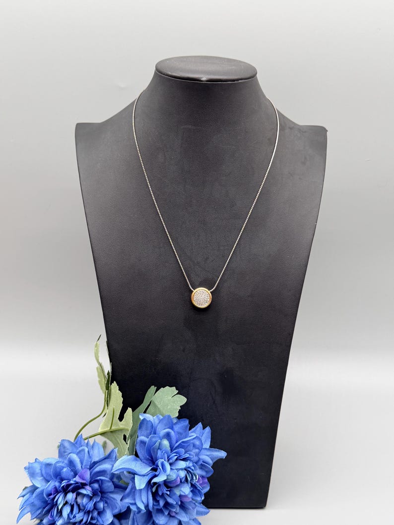 Puede incluir: Un collar plateado con un colgante redondo. El colgante tiene un borde dorado y un centro lleno de peque&ntilde;as piedras brillantes. El collar se exhibe en un maniqu&iacute; negro sobre un fondo blanco, con flores azules en primer plano.