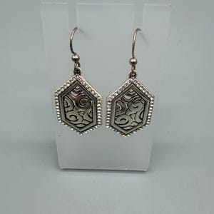 Puede incluir: Un par de pendientes colgantes plateados. Cada pendiente presenta una forma hexagonal con un patr&oacute;n de remolino decorativo en el centro, enmarcado por peque&ntilde;os cristales transparentes. Los pendientes cuelgan de un gancho simple.