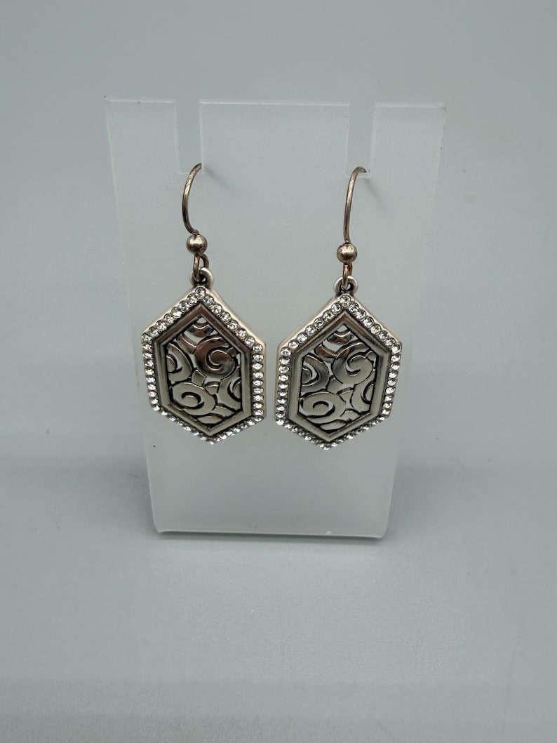 Puede incluir: Un par de pendientes colgantes plateados. Cada pendiente presenta una forma hexagonal con un patr&oacute;n de remolino decorativo en el centro, enmarcado por peque&ntilde;os cristales transparentes. Los pendientes cuelgan de un gancho simple.