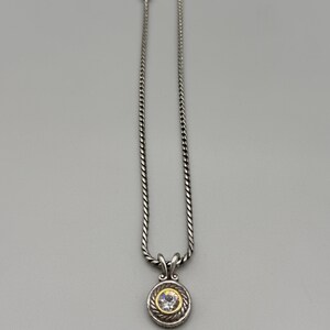 May include: A silver-toned necklace featuring a twisted rope chain and a circular pendant. The pendant has a central clear round stone set within a gold-toned border, surrounded by a textured silver-toned rope frame and a decorative bail.