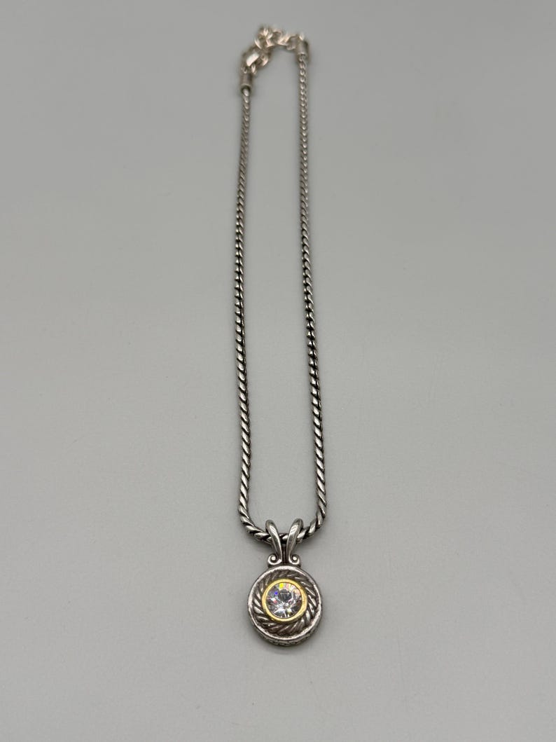 May include: A silver-toned necklace featuring a twisted rope chain and a circular pendant. The pendant has a central clear round stone set within a gold-toned border, surrounded by a textured silver-toned rope frame and a decorative bail.