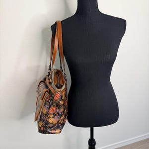 May include: A floral print handbag with brown leather straps and accents. The bag features a colorful pattern of roses and other flowers against a dark background. The bag is displayed on a black mannequin.