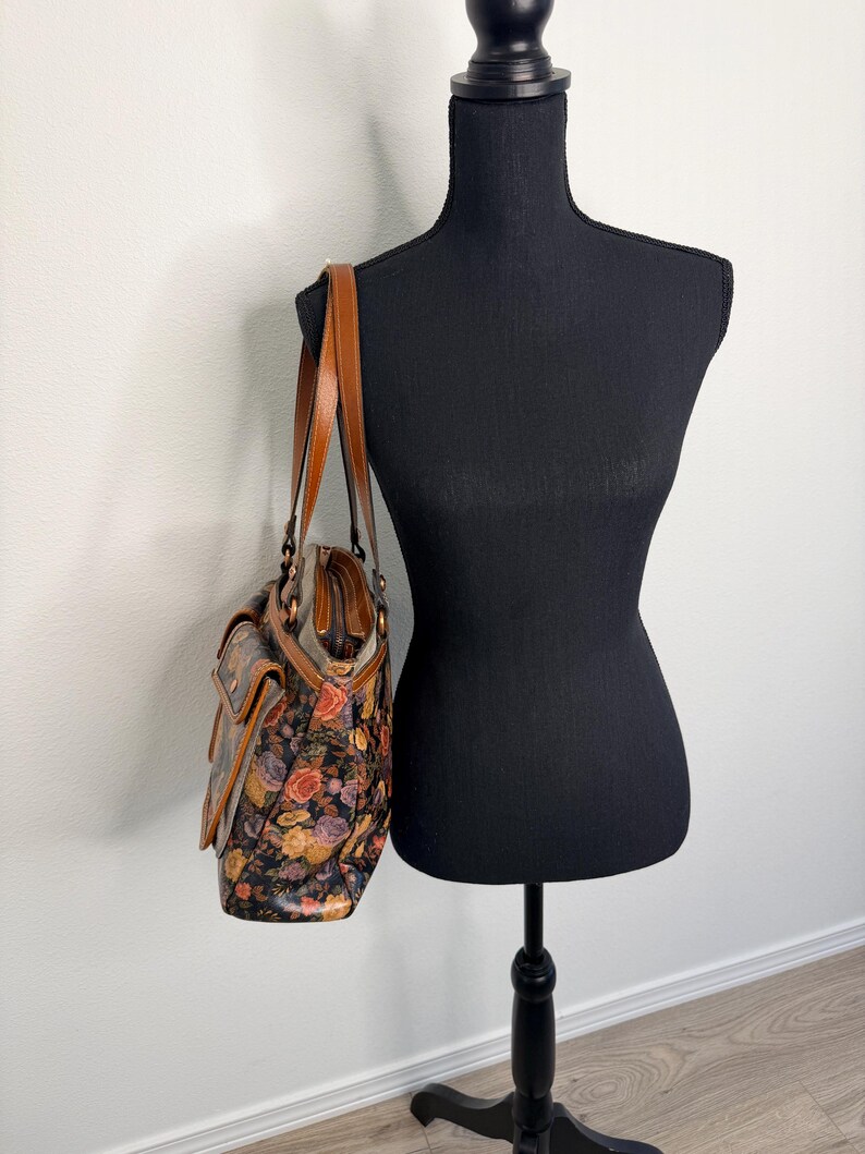 May include: A floral print handbag with brown leather straps and accents. The bag features a colorful pattern of roses and other flowers against a dark background. The bag is displayed on a black mannequin.
