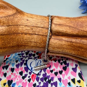 May include: A silver chain bracelet featuring a small, detailed lipstick charm with a pink tip. The charm is attached to a woven metal link chain. The bracelet is displayed on a wooden stand, positioned above a white fabric pouch covered in colourful hearts.