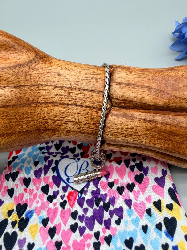 May include: A silver chain bracelet featuring a small, detailed lipstick charm with a pink tip. The charm is attached to a woven metal link chain. The bracelet is displayed on a wooden stand, positioned above a white fabric pouch covered in colourful hearts.