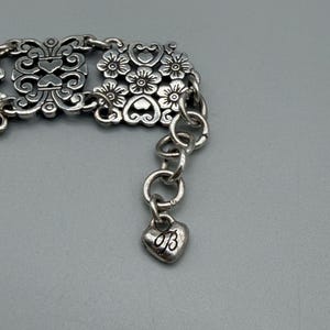以下が含まれることがあります： 銀色のブレスレット。装飾的な花柄とスクロールワークのデザイン。ブレスレットには、文字「B」のハート型のチャームが付いています。ブレスレットは金属製で、チェーンエクステンションが付いています。