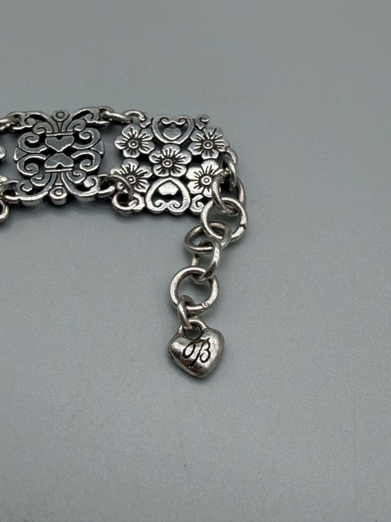 以下が含まれることがあります： 銀色のブレスレット。装飾的な花柄とスクロールワークのデザイン。ブレスレットには、文字「B」のハート型のチャームが付いています。ブレスレットは金属製で、チェーンエクステンションが付いています。