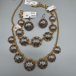 May include: A gold-toned necklace and matching earrings set. The jewelry features silver-colored, circular pendants with intricate designs. The necklace and earrings are displayed on a light gray surface. The tags read "ART OF GOLD".