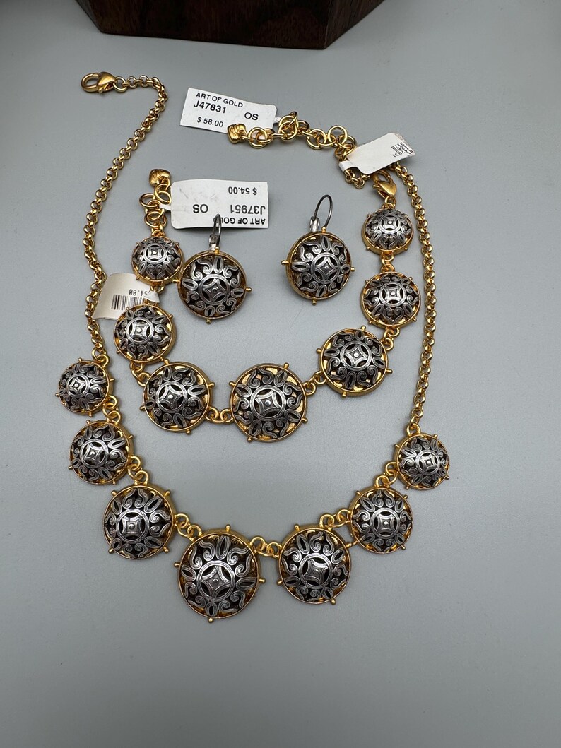 May include: A gold-toned necklace and matching earrings set. The jewelry features silver-colored, circular pendants with intricate designs. The necklace and earrings are displayed on a light gray surface. The tags read "ART OF GOLD".