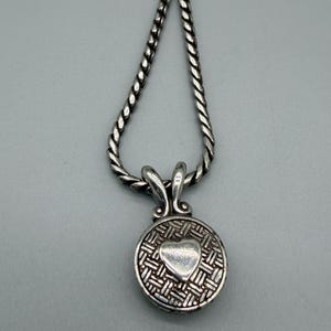 May include: A silver-toned necklace featuring a twisted rope chain and a circular pendant. The pendant displays a textured, woven-style background with a smooth, raised heart shape in the center, attached to the chain by a decorative bail.