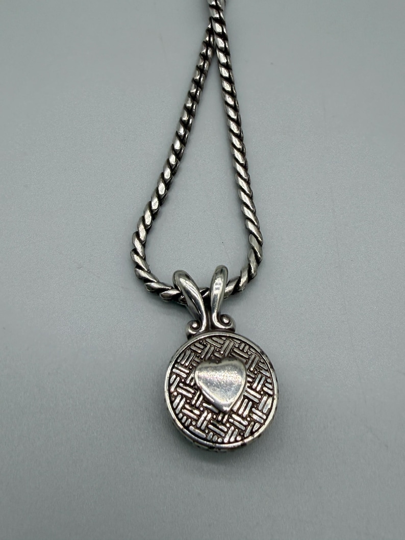 May include: A silver-toned necklace featuring a twisted rope chain and a circular pendant. The pendant displays a textured, woven-style background with a smooth, raised heart shape in the center, attached to the chain by a decorative bail.