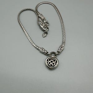 Puede incluir: Collar plateado con un colgante en forma de coraz&oacute;n con un dise&ntilde;o intrincado. El collar tiene una cadena trenzada y un cierre de mosquet&oacute;n. El colgante est&aacute; detallado con un dise&ntilde;o de nudo celta. El collar se muestra sobre un fondo gris.