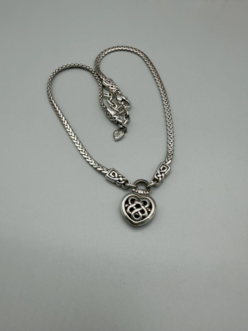 Puede incluir: Collar plateado con un colgante en forma de coraz&oacute;n con un dise&ntilde;o intrincado. El collar tiene una cadena trenzada y un cierre de mosquet&oacute;n. El colgante est&aacute; detallado con un dise&ntilde;o de nudo celta. El collar se muestra sobre un fondo gris.