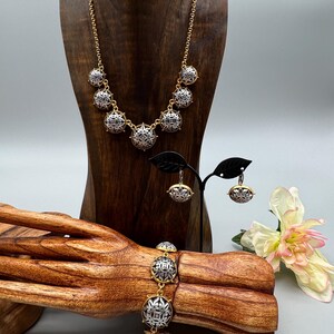 May include: A gold-toned necklace, bracelet, and earrings set. The jewelry features silver and gold circular pendants with an intricate design. The jewelry is displayed on a wooden bust and a wooden hand-shaped stand.