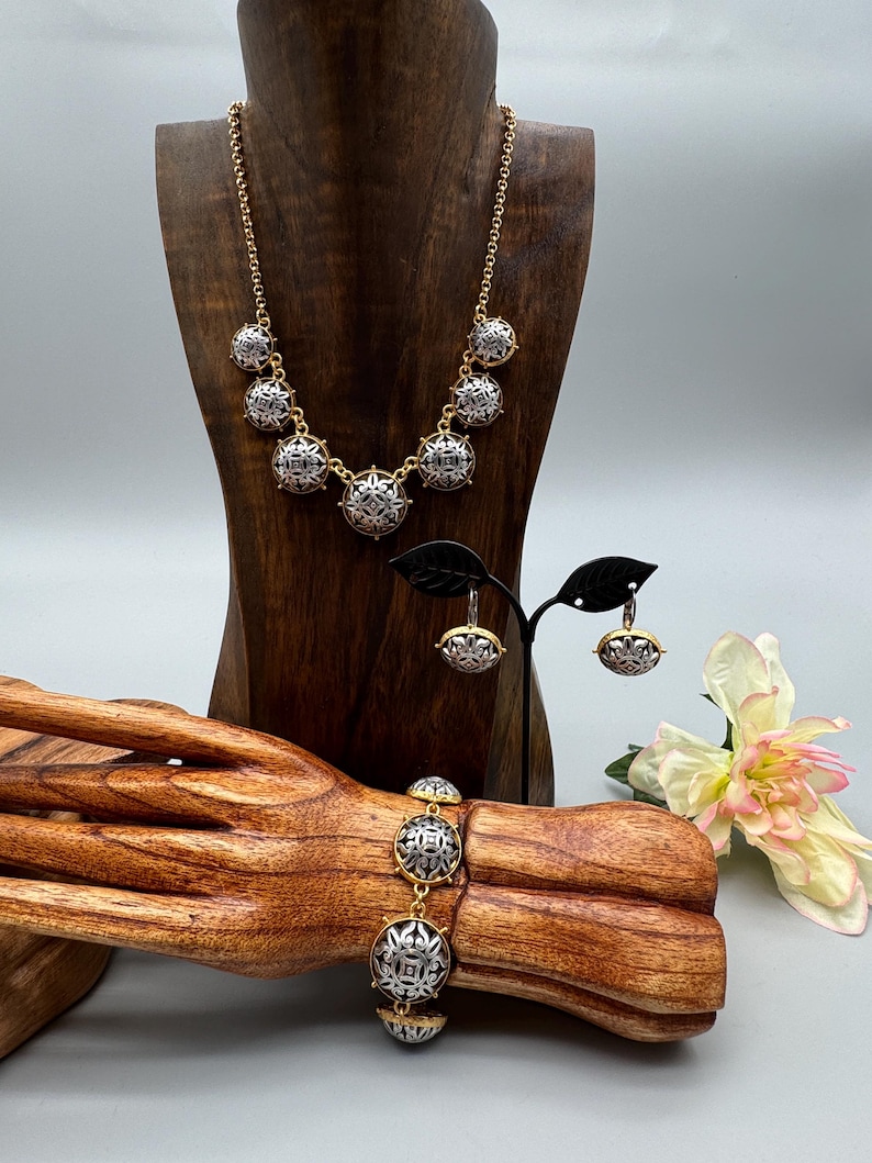 May include: A gold-toned necklace, bracelet, and earrings set. The jewelry features silver and gold circular pendants with an intricate design. The jewelry is displayed on a wooden bust and a wooden hand-shaped stand.