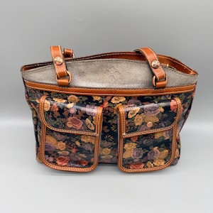 May include: A floral print tote bag with two front pockets. The bag features brown leather handles and trim, with a gray top section. The floral pattern includes roses in shades of pink, yellow, and purple against a dark background.