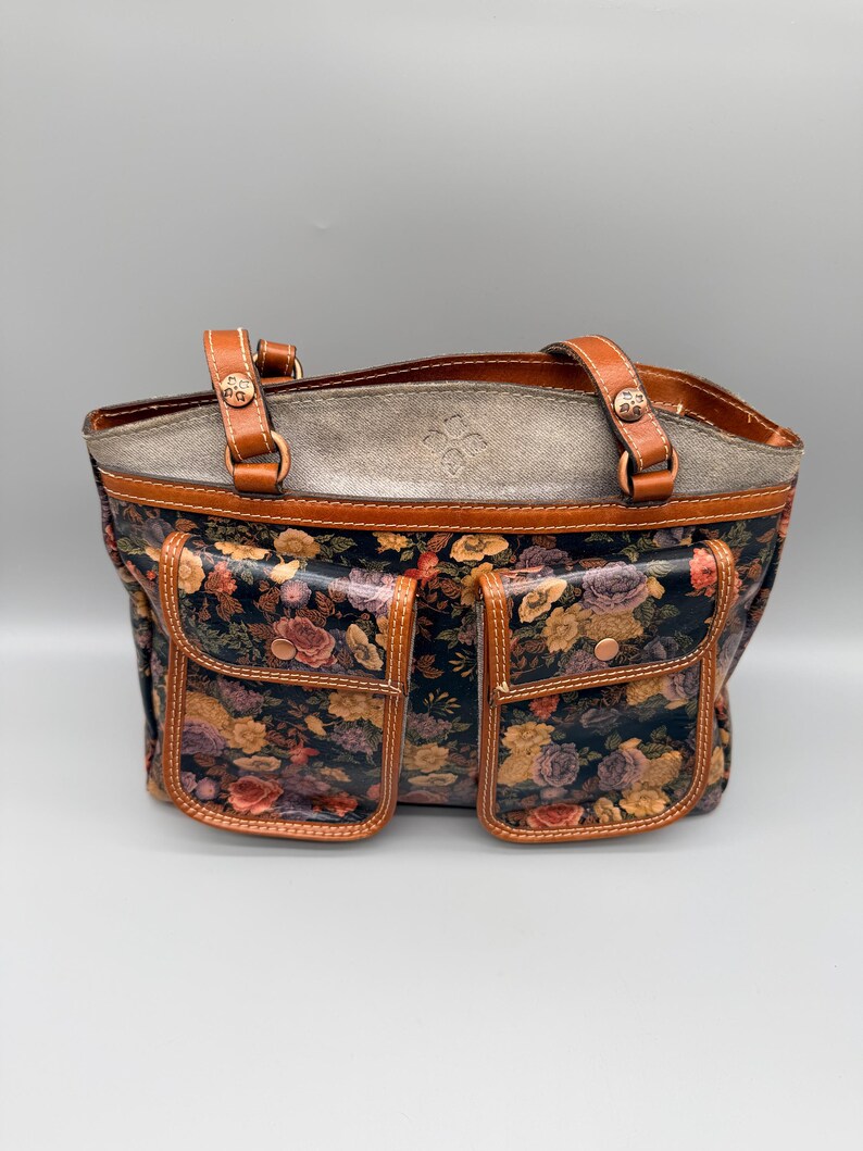 May include: A floral print tote bag with two front pockets. The bag features brown leather handles and trim, with a gray top section. The floral pattern includes roses in shades of pink, yellow, and purple against a dark background.