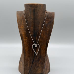 Pu&ograve; includere: Una collana con pendente a cuore argentato esposta su un busto di legno per gioielli. Il pendente a forma di cuore &egrave; delineato con piccole pietre scintillanti. La catena della collana &egrave; delicata e argentata.