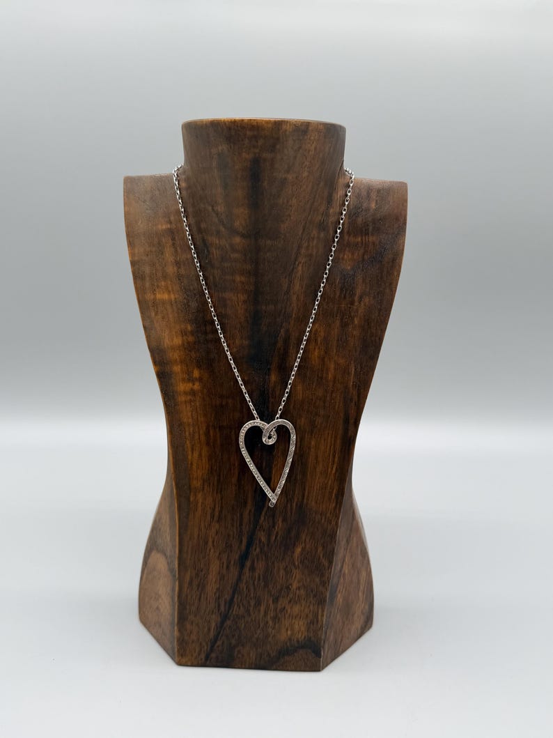 Pu&ograve; includere: Una collana con pendente a cuore argentato esposta su un busto di legno per gioielli. Il pendente a forma di cuore &egrave; delineato con piccole pietre scintillanti. La catena della collana &egrave; delicata e argentata.