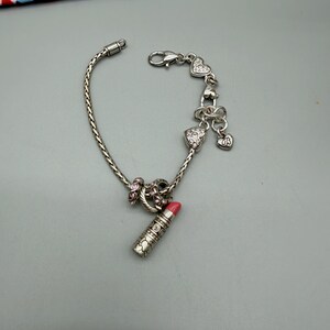 May include: A silver-tone charm bracelet featuring a braided chain, a lipstick charm with a pink tip, and several heart-shaped charms adorned with clear crystals. The bracelet includes a lobster claw clasp and a dangling heart charm with the letters US.