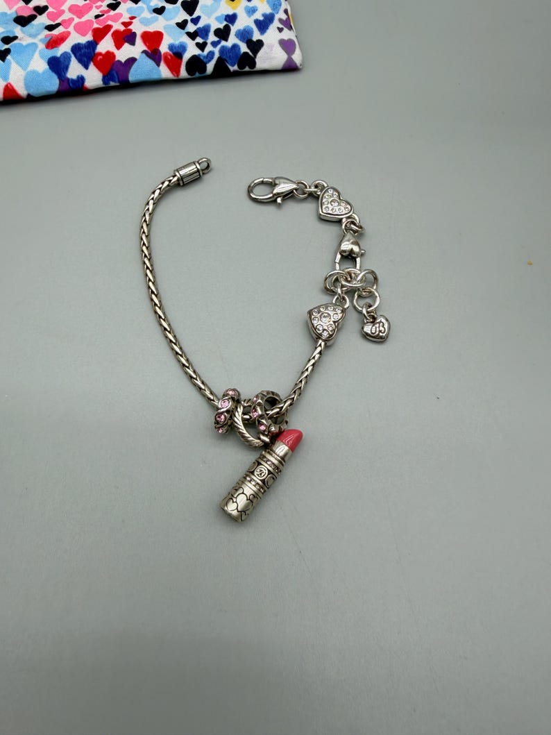 May include: A silver-tone charm bracelet featuring a braided chain, a lipstick charm with a pink tip, and several heart-shaped charms adorned with clear crystals. The bracelet includes a lobster claw clasp and a dangling heart charm with the letters US.