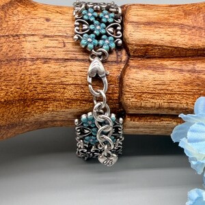 以下が含まれることがあります： ターコイズブルーの花柄が特徴のシルバーブレスレット。ブレスレットは、精巧なデザインとターコイズブルーの花が施された2つの長方形のセクションで構成されています。セクションはハート型のリンクのチェーンで接続されています。「OB」のイニシャルが入った小さなチャームが見えます。
