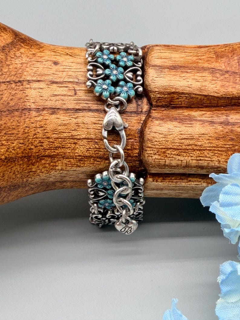 以下が含まれることがあります： ターコイズブルーの花柄が特徴のシルバーブレスレット。ブレスレットは、精巧なデザインとターコイズブルーの花が施された2つの長方形のセクションで構成されています。セクションはハート型のリンクのチェーンで接続されています。「OB」のイニシャルが入った小さなチャームが見えます。