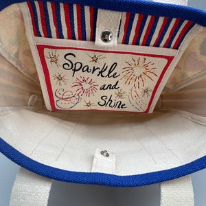Puede incluir: Un bolso de mano de lona con ribete y correas azules. El interior presenta un bolsillo con las palabras "Sparkle and Shine" y fuegos artificiales. El bolsillo tiene un borde a rayas rojas y blancas.