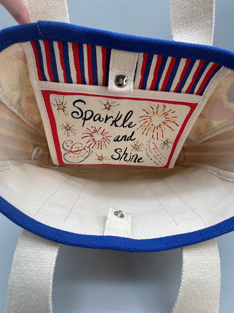 Puede incluir: Un bolso de mano de lona con ribete y correas azules. El interior presenta un bolsillo con las palabras "Sparkle and Shine" y fuegos artificiales. El bolsillo tiene un borde a rayas rojas y blancas.