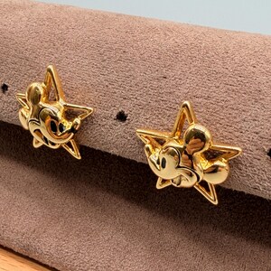 May include: A pair of gold-colored star-shaped earrings. Each earring features a Mickey Mouse design in the center. The earrings are displayed on a brown surface.