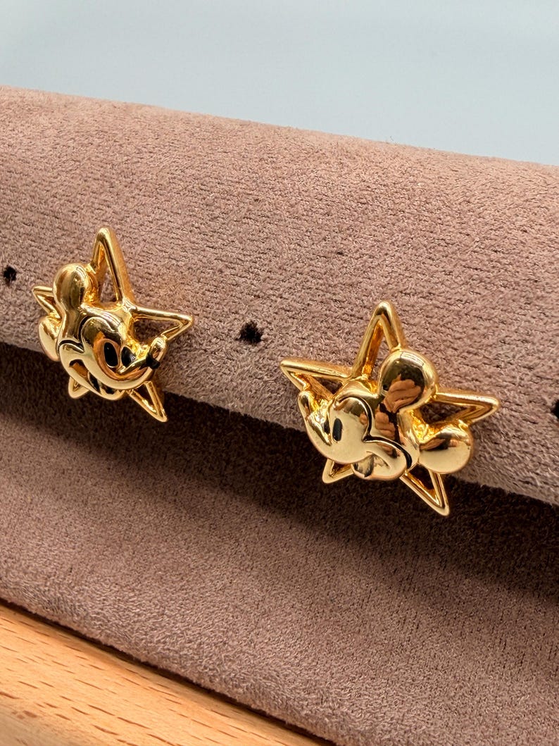 May include: A pair of gold-colored star-shaped earrings. Each earring features a Mickey Mouse design in the center. The earrings are displayed on a brown surface.