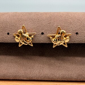 May include: A pair of gold-colored star-shaped stud earrings. Each earring features a Mickey Mouse design in the center. The earrings are displayed on a brown surface.