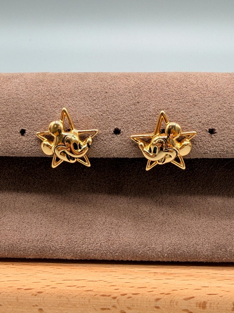 May include: A pair of gold-colored star-shaped stud earrings. Each earring features a Mickey Mouse design in the center. The earrings are displayed on a brown surface.
