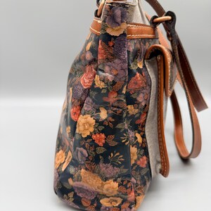 May include: A floral-patterned handbag with a dark background and colorful flowers in shades of orange, purple, and yellow. The bag has brown leather straps and trim, with a neutral-colored fabric on the sides.