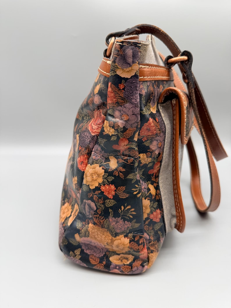 May include: A floral-patterned handbag with a dark background and colorful flowers in shades of orange, purple, and yellow. The bag has brown leather straps and trim, with a neutral-colored fabric on the sides.