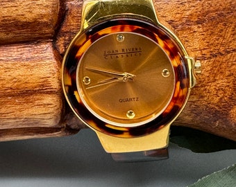 May include: A Joan Rivers Classics quartz watch featuring a round gold-tone case and a brown tortoiseshell-patterned bezel and band. The watch face is a metallic bronze color with gold-tone hands, four small circular hour markers, and the text.
