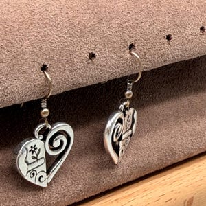 May include: A pair of silver heart-shaped earrings. Each earring has a half-heart design with a floral motif and a swirling scroll. The earrings hang from a simple hook.