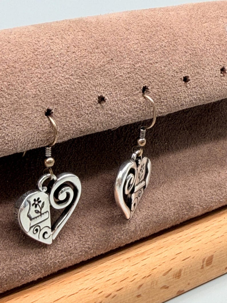 May include: A pair of silver heart-shaped earrings. Each earring has a half-heart design with a floral motif and a swirling scroll. The earrings hang from a simple hook.