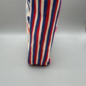 Puede incluir: Un recipiente de tela cil&iacute;ndrico y alto, con borde y base azules. El recipiente presenta rayas verticales rojas, blancas y azules. El lateral tiene formas abstractas coloridas.