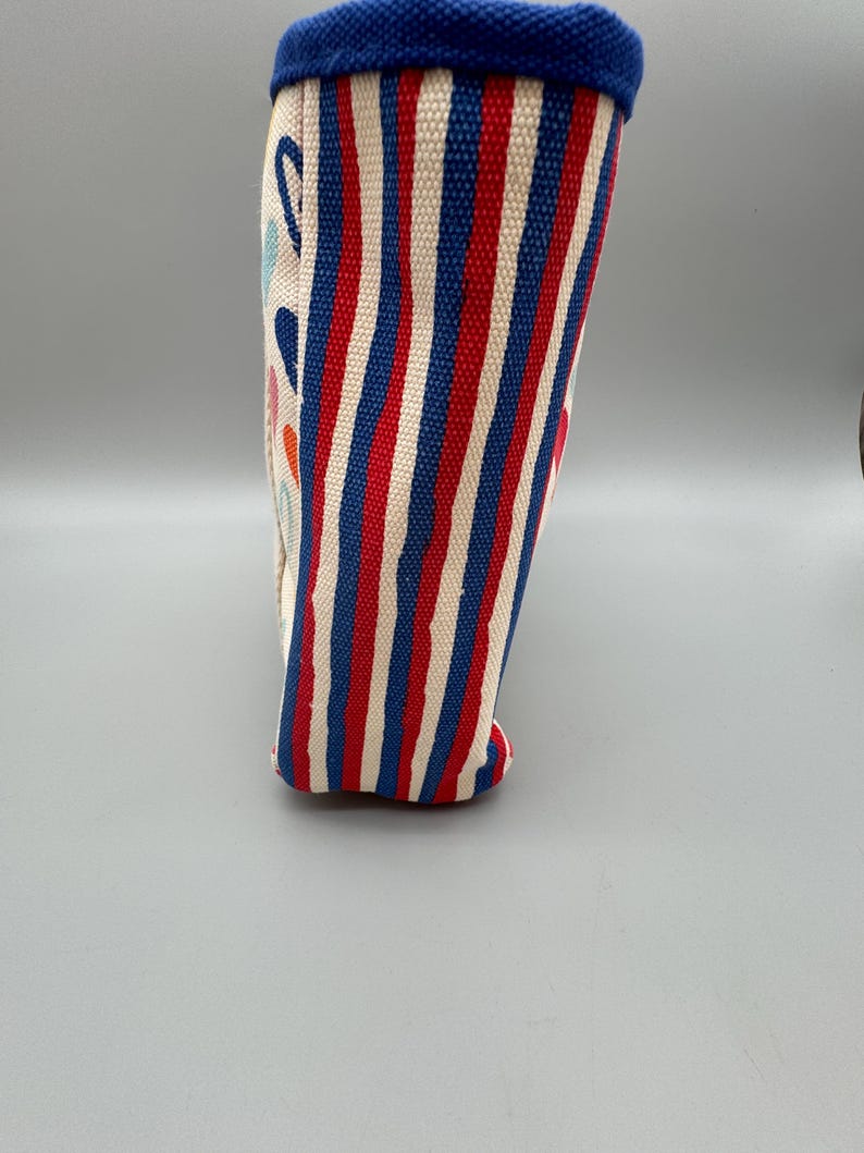 Puede incluir: Un recipiente de tela cil&iacute;ndrico y alto, con borde y base azules. El recipiente presenta rayas verticales rojas, blancas y azules. El lateral tiene formas abstractas coloridas.