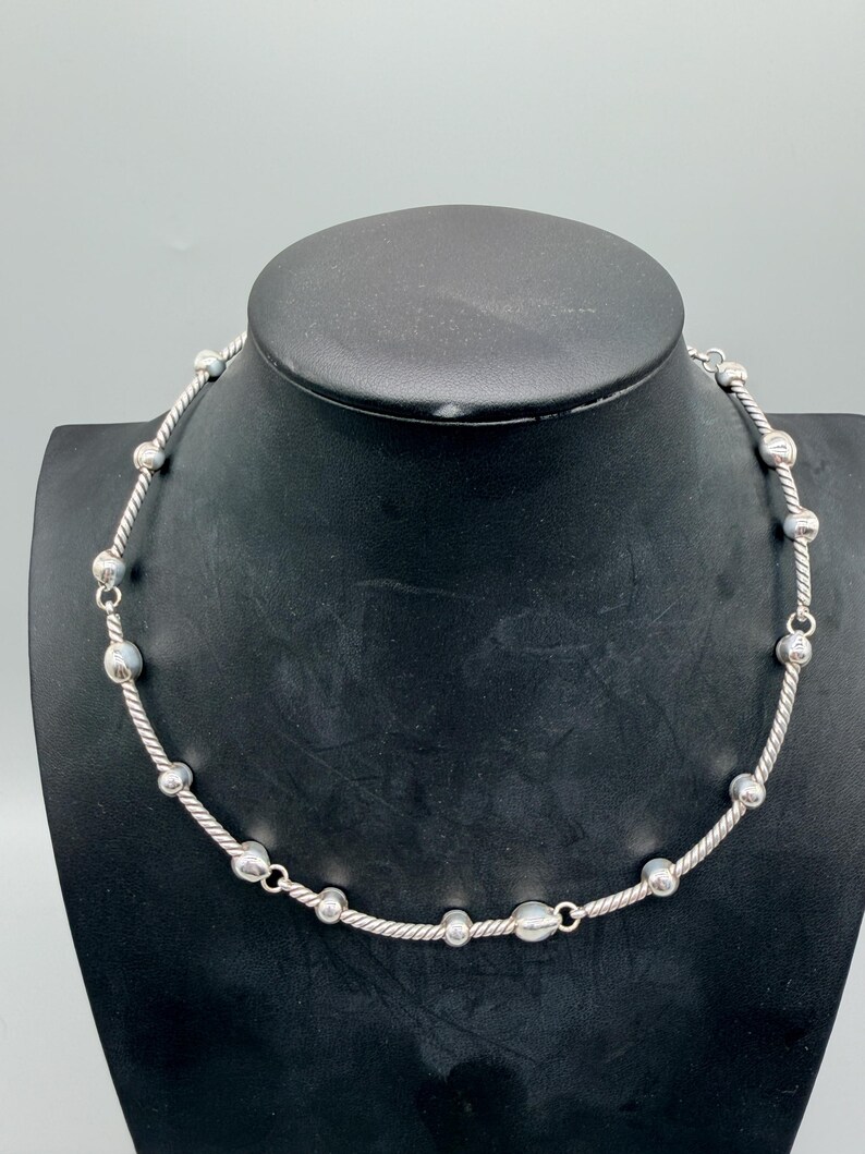 May include: A silver-toned necklace featuring a series of smooth, round metallic beads interspersed along a twisted, rope-textured chain. The design alternates between the polished spherical beads and segments of the twisted metal wire throughout.