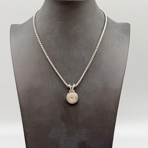 May include: A silver chain necklace featuring a circular pendant with a central clear stone, surrounded by a gold-toned inner ring and a textured silver outer border. The pendant hangs from a silver bail on a twisted rope-style silver chain.