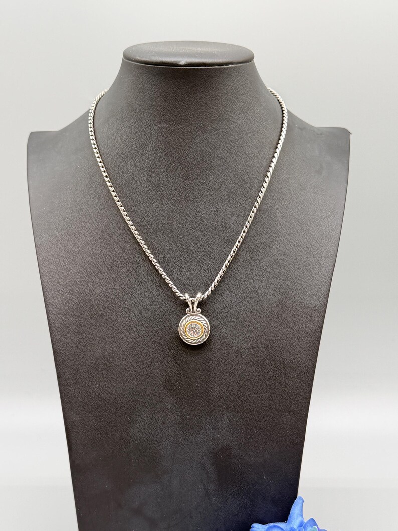 May include: A silver chain necklace featuring a circular pendant with a central clear stone, surrounded by a gold-toned inner ring and a textured silver outer border. The pendant hangs from a silver bail on a twisted rope-style silver chain.