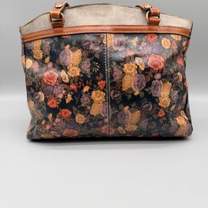 May include: A floral print tote bag with a black background, featuring roses in shades of orange, yellow, and purple. The bag has brown leather handles and trim, with a light gray top section.