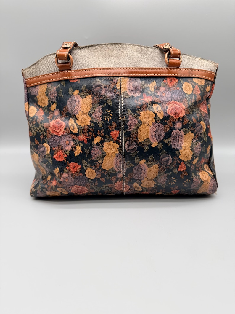 May include: A floral print tote bag with a black background, featuring roses in shades of orange, yellow, and purple. The bag has brown leather handles and trim, with a light gray top section.