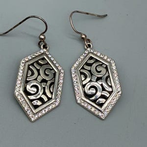 Puede incluir: Un par de pendientes colgantes plateados. Cada pendiente presenta un dise&ntilde;o geom&eacute;trico calado con un patr&oacute;n de remolino, enmarcado por un borde de peque&ntilde;os cristales transparentes. Cierre de gancho.